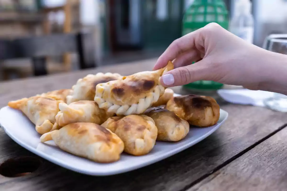 A hand picking up an empanada from a plate with several empanadas on a wooden table.