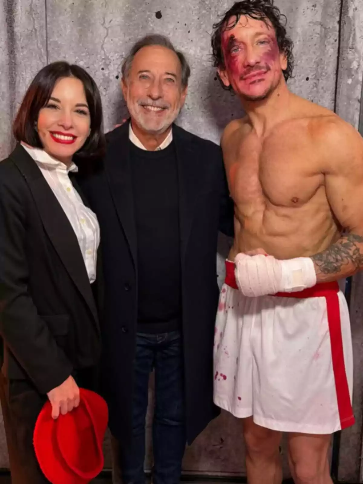 Three people pose smiling in front of a gray wall: a woman holding a red hat, an older man with a beard, and a jerseyless man with a bandage on his hand and makeup simulating wounds on his face and torso. Three people pose smiling in front of a gray wall: a woman holding a red hat, an older man with a beard, and a jerseyless man with a bandage on his hand and makeup simulating wounds on his face and torso.