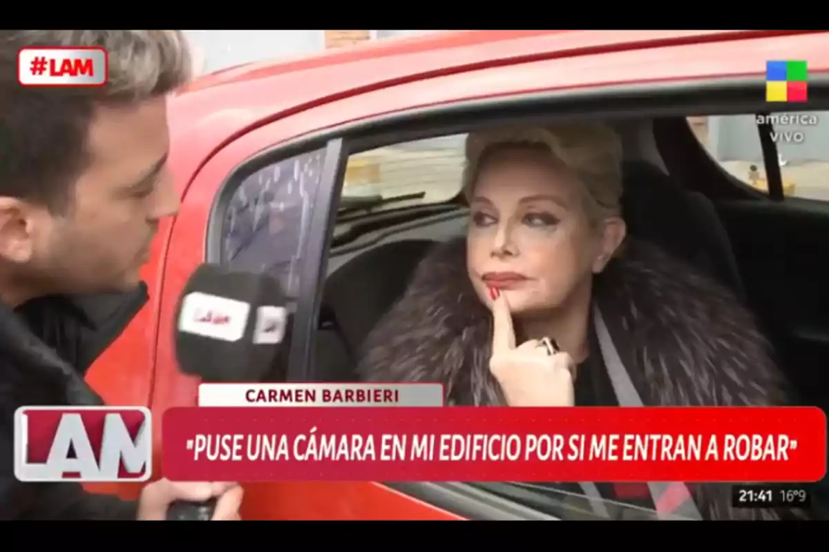 Una mujer sentada en un auto rojo es entrevistada por un periodista que sostiene un micrófono mientras ella apoya un dedo en su barbilla y mira pensativa Una mujer sentada en un auto rojo es entrevistada por un periodista que sostiene un micrófono mientras ella apoya un dedo en su barbilla y mira pensativa