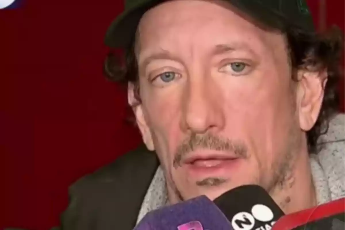 Hombre de cabello rizado y gorra negra hablando frente a varios micrófonos Hombre de cabello rizado y gorra negra hablando frente a varios micrófonos