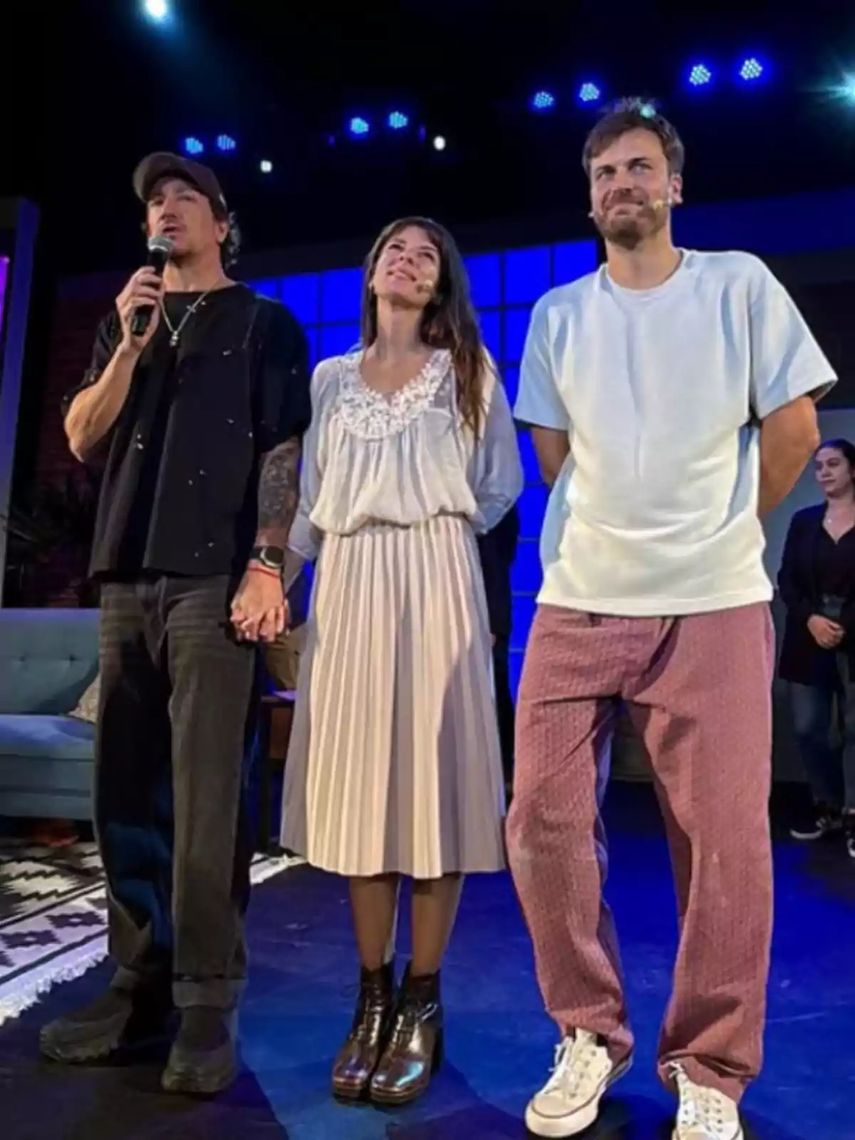 Tres personas de pie en un escenario iluminado, una de ellas sostiene un micrófono y otra persona está en el centro sonriendo mientras se toman de la mano Tres personas de pie en un escenario iluminado, una de ellas sostiene un micrófono y otra persona está en el centro sonriendo mientras se toman de la mano