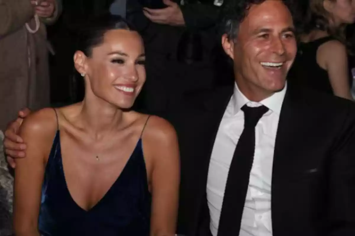 Una pareja elegantemente vestida sonríe mientras está sentada en un evento social Una pareja elegantemente vestida sonríe mientras está sentada en un evento social