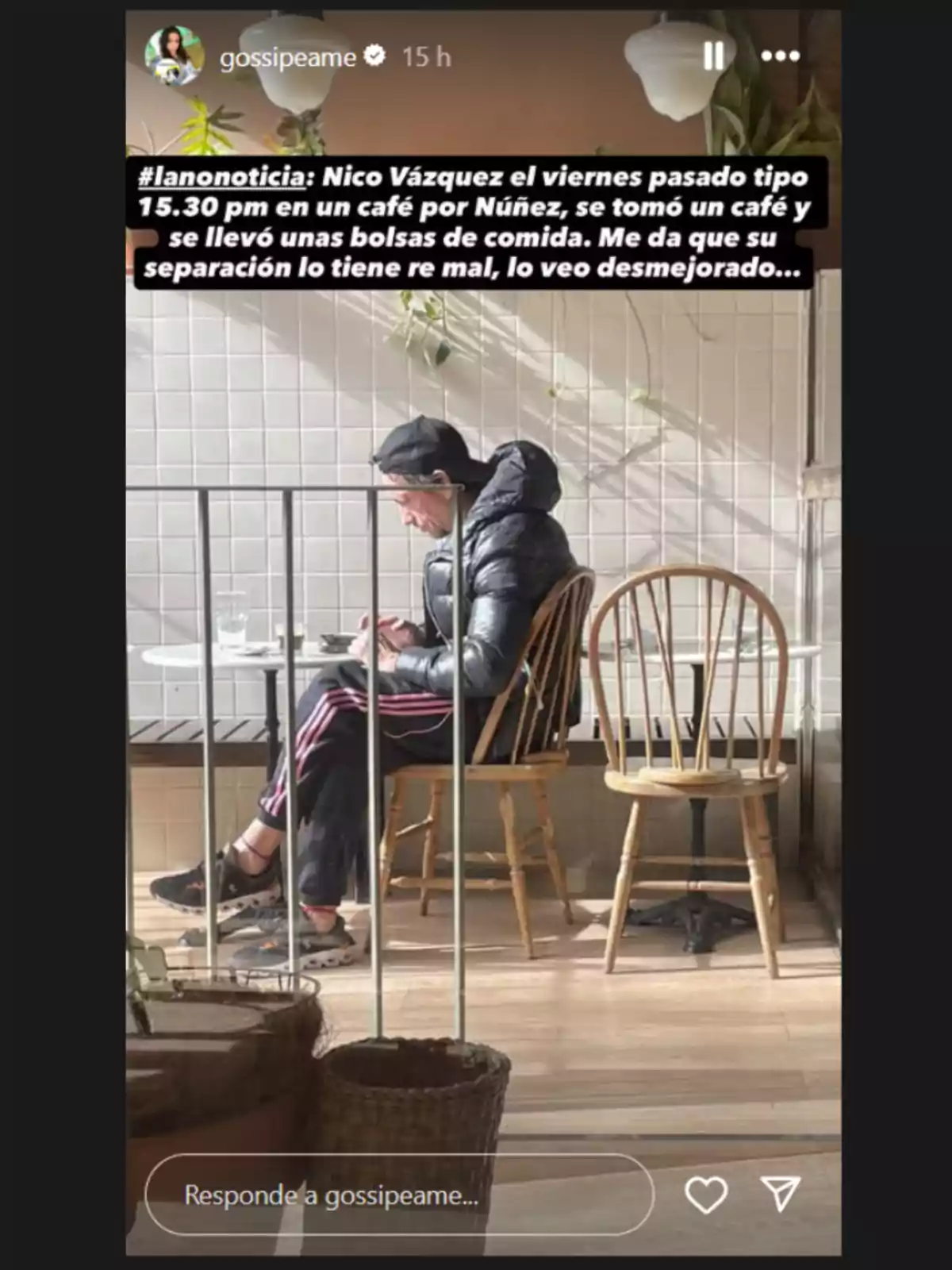 Un hombre con gorra negra y campera inflable está sentado solo en una mesa de un café, usando el celular y con bolsas de comida sobre la mesa, mientras la luz del sol entra por la ventana. Un hombre con gorra negra y campera inflable está sentado solo en una mesa de un café, usando el celular y con bolsas de comida sobre la mesa, mientras la luz del sol entra por la ventana.