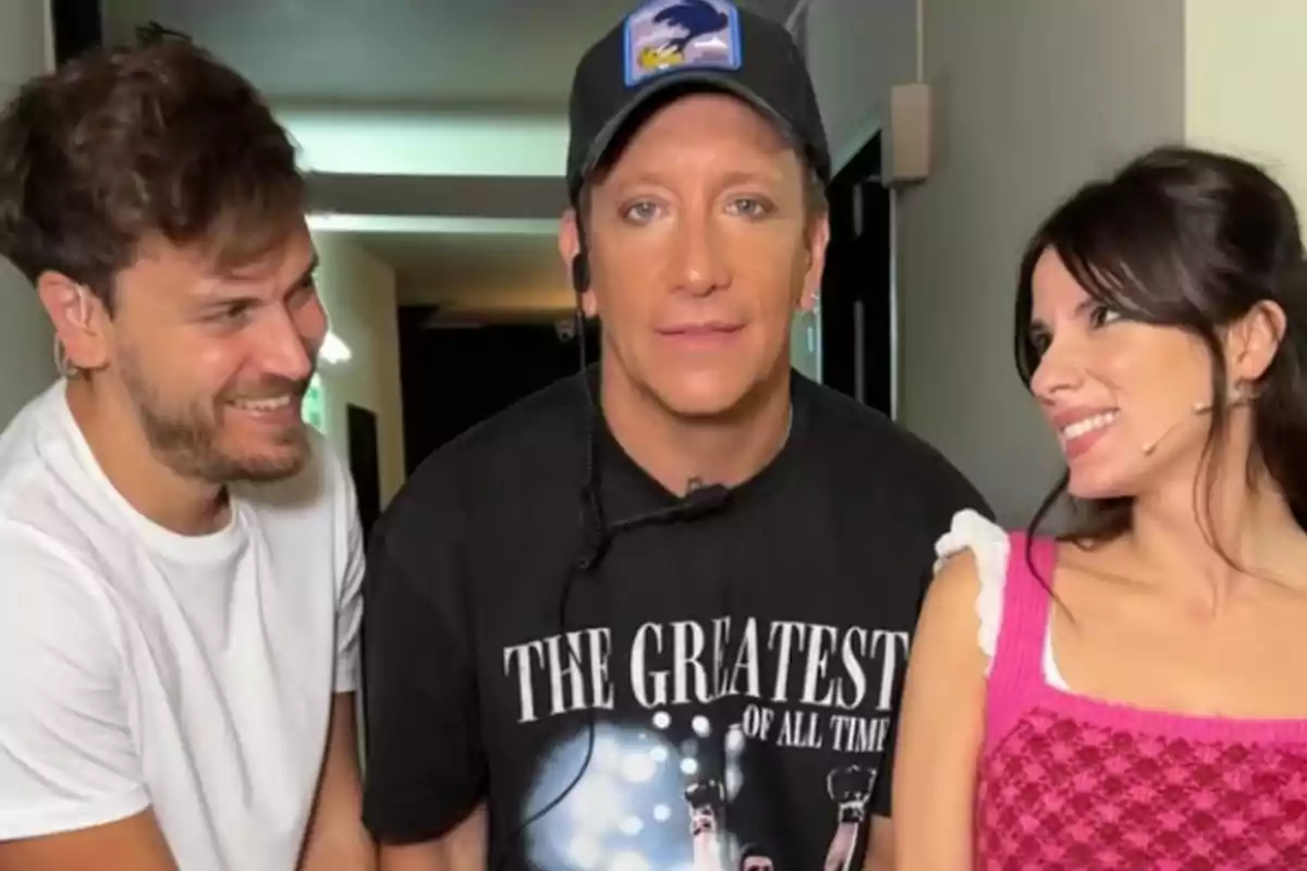 Three people pose together in a hallway; the man in the center is wearing a black cap and a T-jersey with white letters, while the other two smile and look at him. Three people pose together in a hallway; the man in the center is wearing a black cap and a T-jersey with white letters, while the other two smile and look at him.