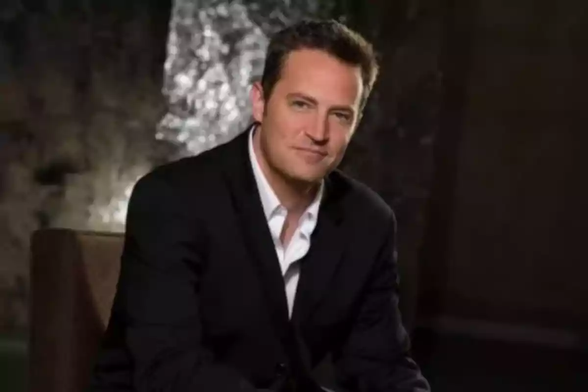 Man with short dark hair wearing a black suit and white jersey sitting on a chair with a dark, blurred background Man with short dark hair wearing a black suit and white jersey sitting on a chair with a dark, blurred background