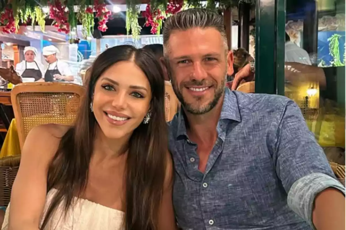 Una pareja sonriente sentada en un restaurante con decoración de flores y chefs trabajando al fondo Una pareja sonriente sentada en un restaurante con decoración de flores y chefs trabajando al fondo
