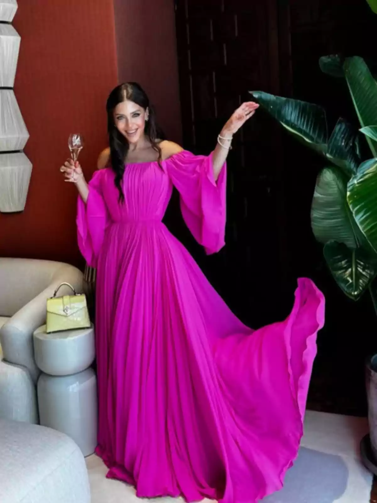 Mujer sonriente con vestido largo fucsia de hombros descubiertos sosteniendo una copa en una mano y levantando la otra, junto a un sillón claro, una bolsa amarilla y una planta grande Mujer sonriente con vestido largo fucsia de hombros descubiertos sosteniendo una copa en una mano y levantando la otra, junto a un sillón claro, una bolsa amarilla y una planta grande