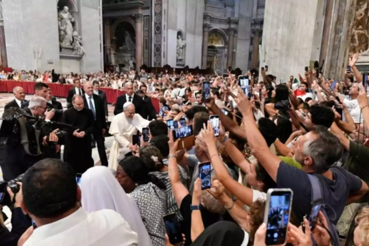 Una multitud de personas dentro de una iglesia levantan sus teléfonos móviles para tomar fotos mientras una figura religiosa camina entre ellos escoltada por seguridad. Una multitud de personas dentro de una iglesia levantan sus teléfonos móviles para tomar fotos mientras una figura religiosa camina entre ellos escoltada por seguridad.