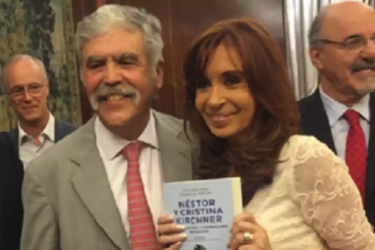 Una mujer con cabello castaño y un hombre de cabello canoso posan sonrientes en un evento mientras ella sostiene un libro azul Una mujer con cabello castaño y un hombre de cabello canoso posan sonrientes en un evento mientras ella sostiene un libro azul