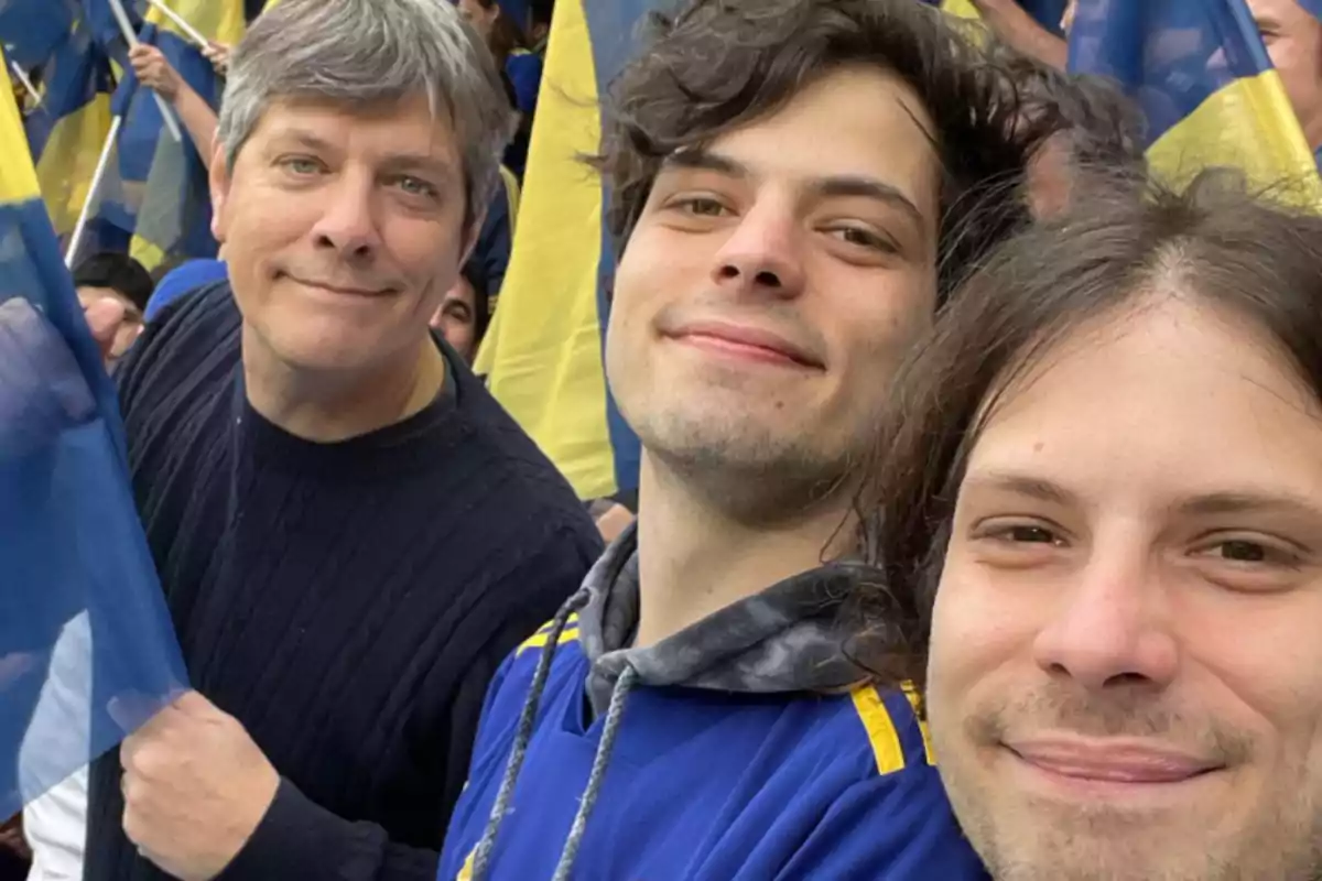 Three people smile as they pose for a selfie in a stadium, surrounded by blue and yellow flags. Three people smile as they pose for a selfie in a stadium, surrounded by blue and yellow flags.