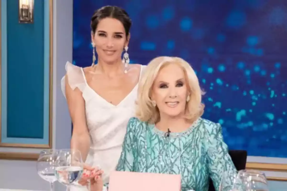 Two elegantly dressed women pose smiling on a television set with glasses of water on the table.