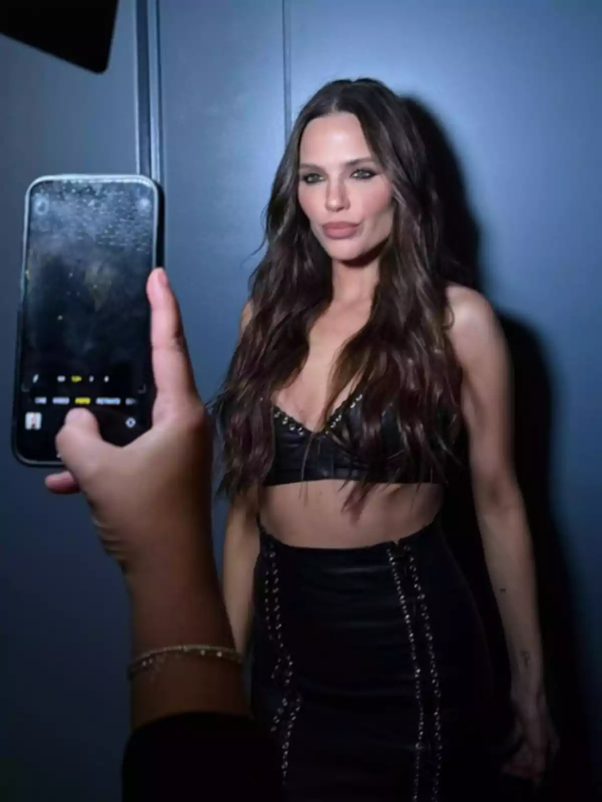 Woman with long, wavy hair poses in front of a blue wall while someone takes her picture with a cellphone. Woman with long, wavy hair poses in front of a blue wall while someone takes her picture with a cellphone.