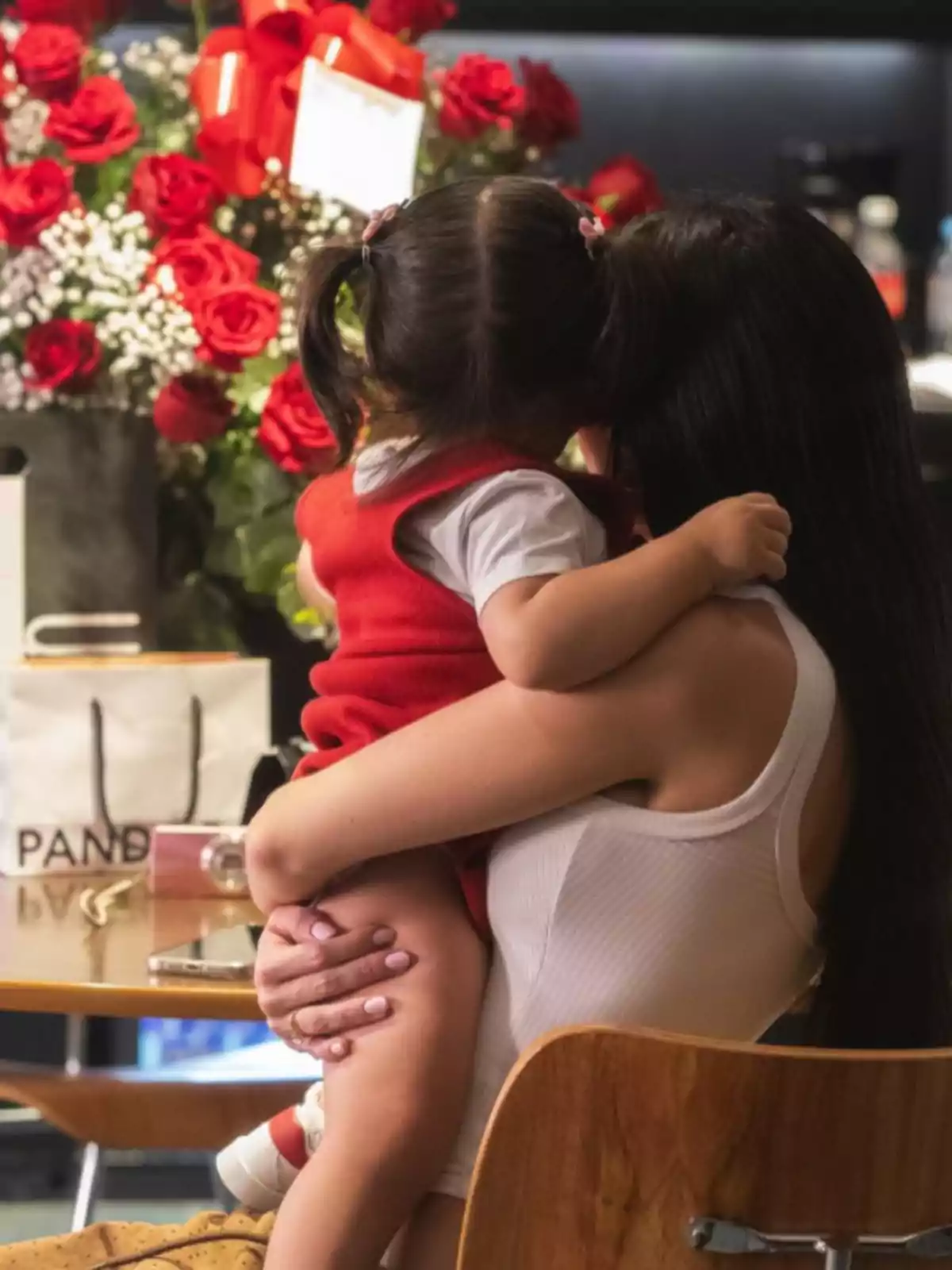Woman sitting, hugging a little girl while they are facing away, with a bouquet of red roses and a gift bag on the table Woman sitting, hugging a little girl while they are facing away, with a bouquet of red roses and a gift bag on the table