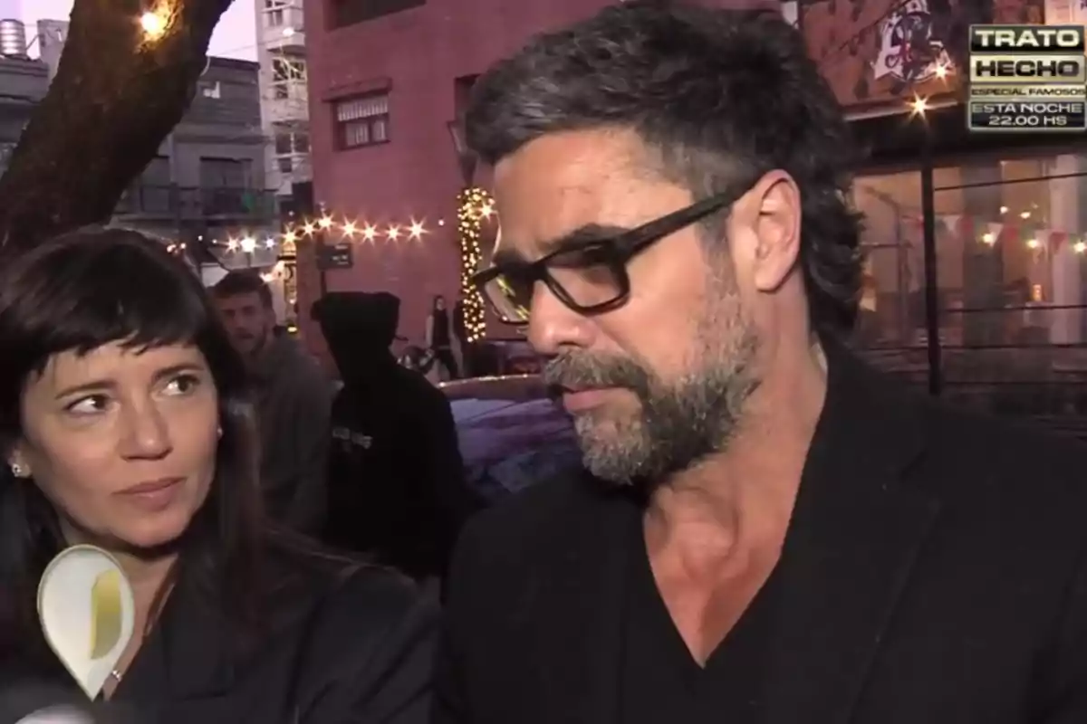 Two people are talking on the street during a nighttime interview with decorative lights in the background and a TV show sign in the top right corner. Two people are talking on the street during a nighttime interview with decorative lights in the background and a TV show sign in the top right corner.