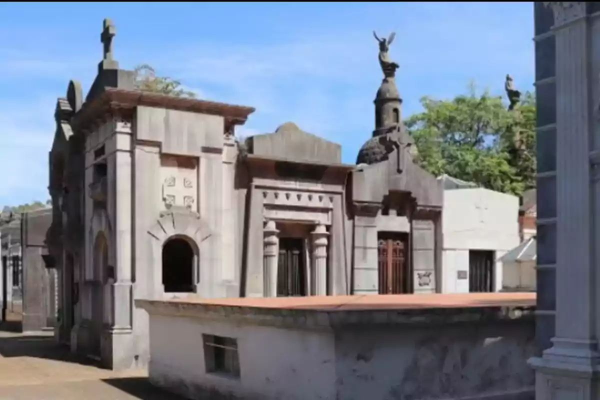 Mausoleos antiguos de piedra en un cementerio con esculturas y árboles al fondo bajo un cielo azul Mausoleos antiguos de piedra en un cementerio con esculturas y árboles al fondo bajo un cielo azul