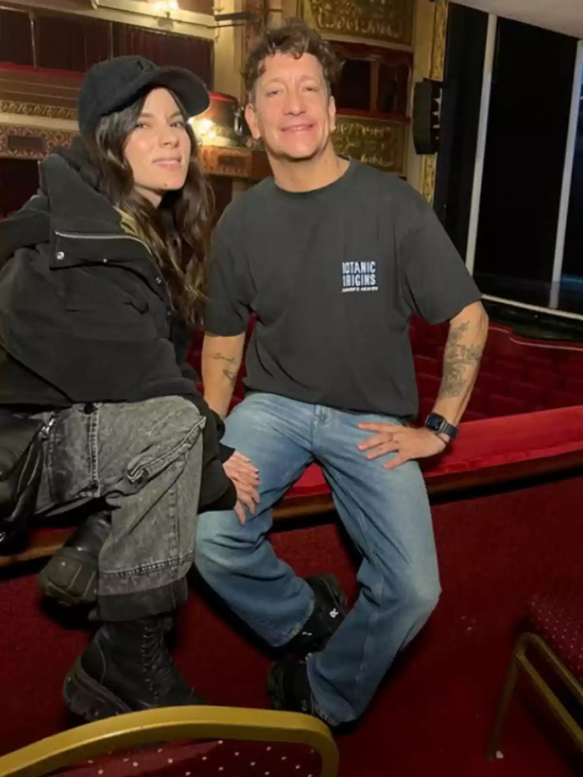 Two people are smiling inside a theater with red seats and golden details; one of them is wearing a cap and dark clothes, while the other is dressed in a black t-jersey and light jeans. Two people are smiling inside a theater with red seats and golden details; one of them is wearing a cap and dark clothes, while the other is dressed in a black t-jersey and light jeans.