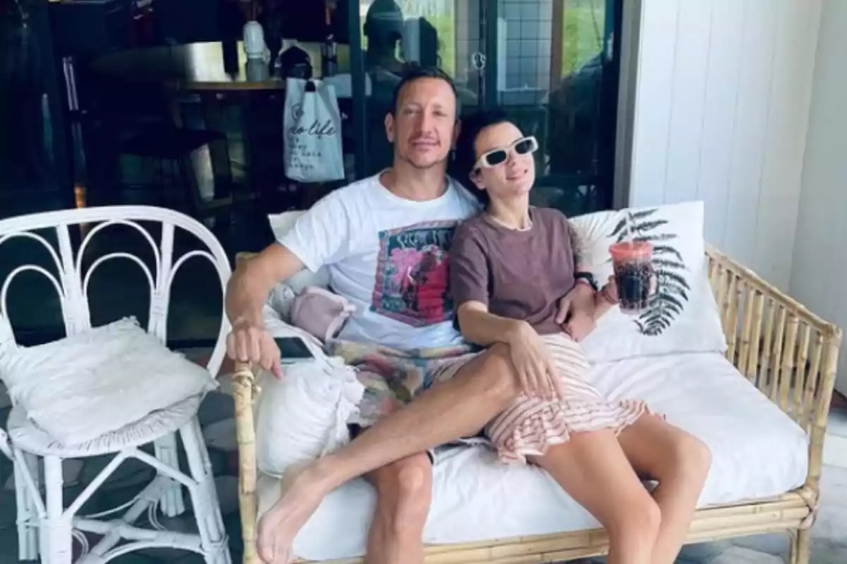 Couple sitting on a wicker sofa on a terrace, hugging and smiling, she is holding a drink with a straw and wearing white sunglasses Couple sitting on a wicker sofa on a terrace, hugging and smiling, she is holding a drink with a straw and wearing white sunglasses