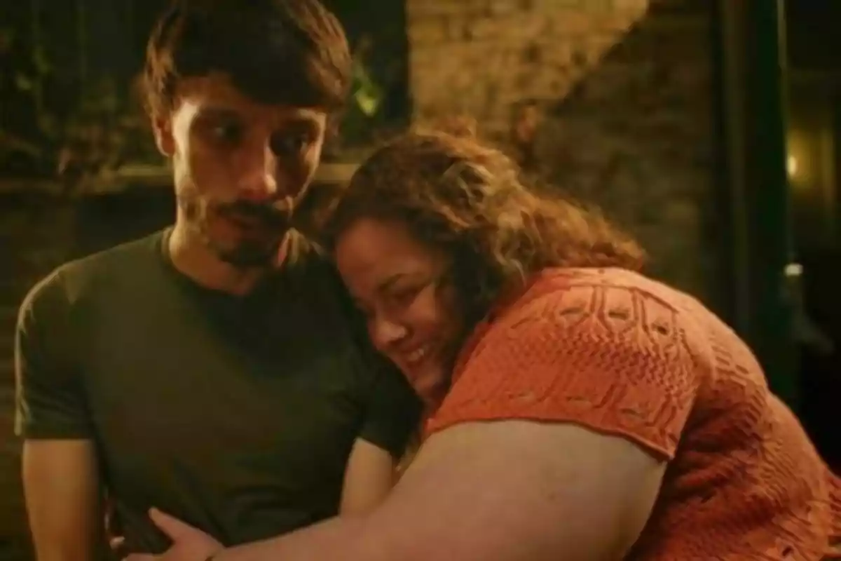 A smiling woman hugs a man who looks uncomfortable in an indoor setting with warm lighting. A smiling woman hugs a man who looks uncomfortable in an indoor setting with warm lighting.