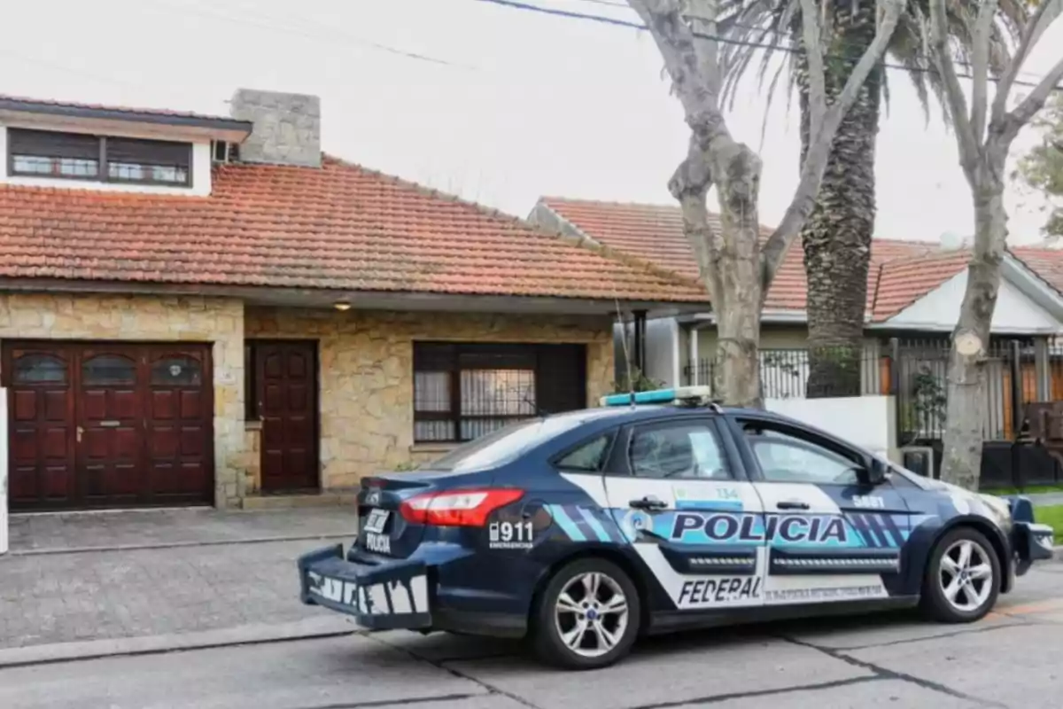Un auto de la policía federal estacionado frente a una casa de fachada de piedra y techo de tejas rojas en una calle arbolada. Un auto de la policía federal estacionado frente a una casa de fachada de piedra y techo de tejas rojas en una calle arbolada.