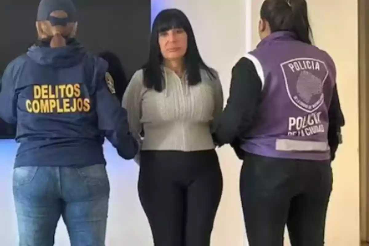 Mujer de cabello oscuro escoltada por dos agentes de policía, una con chaqueta azul que dice delitos complejos y otra con chaleco morado que dice policía de la ciudad Mujer de cabello oscuro escoltada por dos agentes de policía, una con chaqueta azul que dice delitos complejos y otra con chaleco morado que dice policía de la ciudad