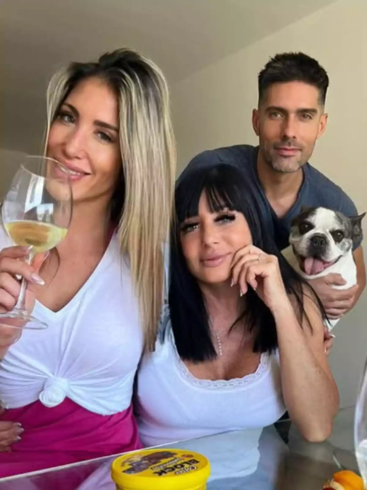Three people smile while posing for a photo at a table; one of them is holding a glass of wine and another is petting a bulldog. There is a yellow container on the table. Three people smile while posing for a photo at a table; one of them is holding a glass of wine and another is petting a bulldog. There is a yellow container on the table.