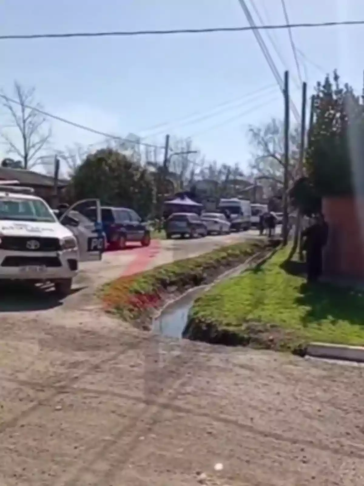 Several police vehicles and a patrol car are parked on a dirt road next to a water-filled ditch in a residential neighborhood during the day. Several police vehicles and a patrol car are parked on a dirt road next to a water-filled ditch in a residential neighborhood during the day.