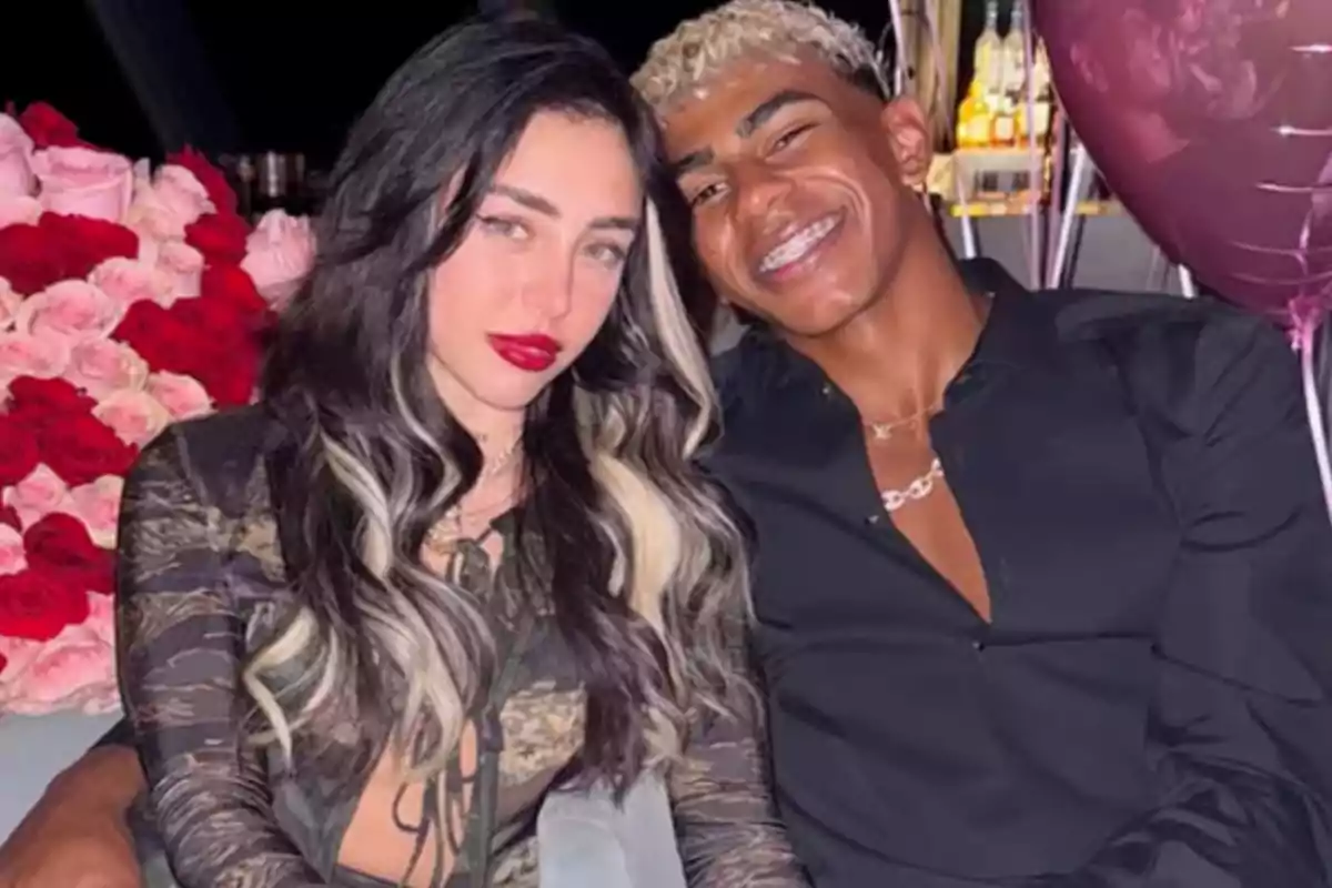 Una pareja joven sonríe sentada en un sofá con globos y un gran ramo de rosas rojas y rosas al fondo Una pareja joven sonríe sentada en un sofá con globos y un gran ramo de rosas rojas y rosas al fondo