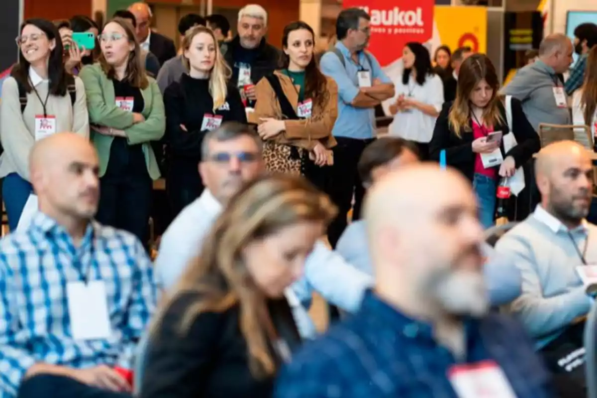 People participating in a business event, some seated and others standing, paying attention or using their mobile phones, in a professional and busy environment.