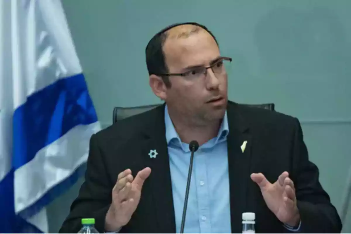 Hombre con gafas y kipá hablando frente a un micrófono con una bandera de Israel al fondo Hombre con gafas y kipá hablando frente a un micrófono con una bandera de Israel al fondo