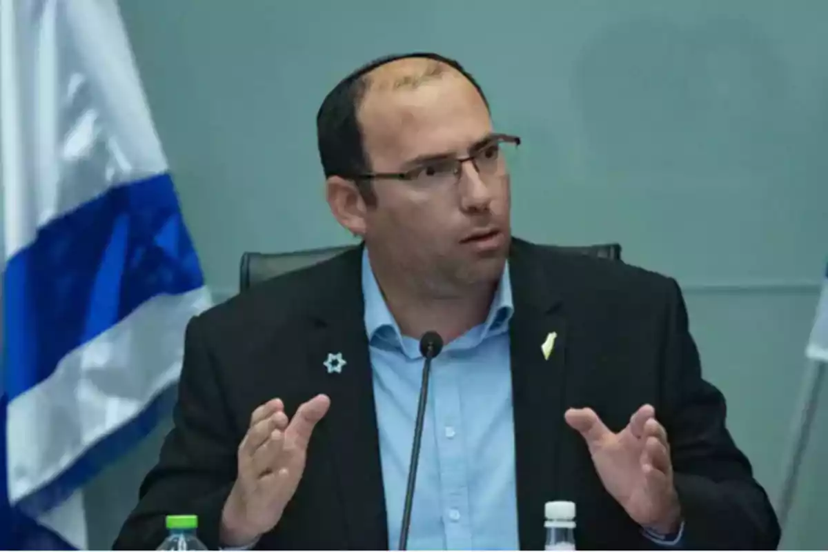 Hombre con gafas y kipá hablando en una conferencia con una bandera de Israel al fondo