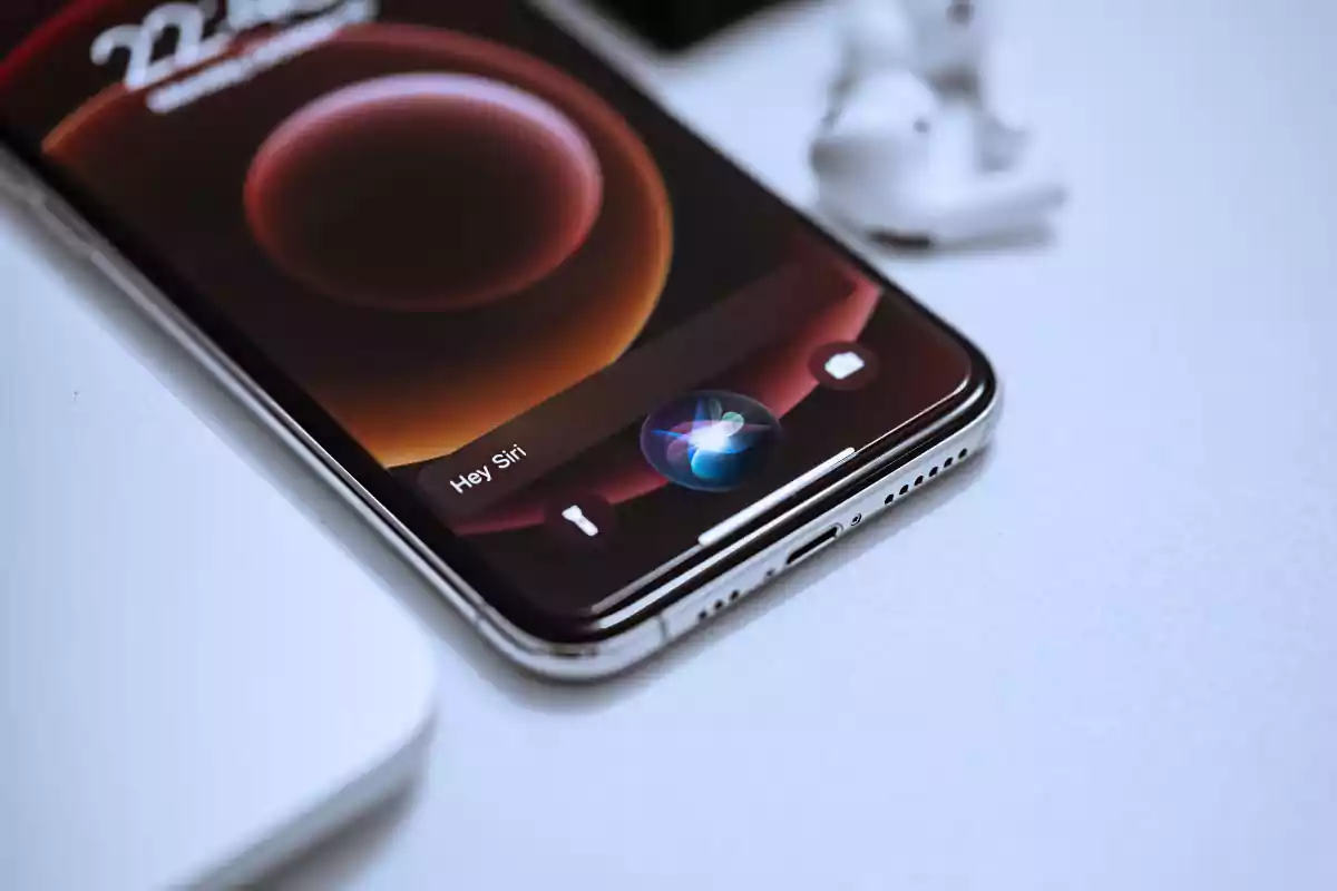 Un iPhone con la pantalla encendida mostrando la interfaz de Siri y unos auriculares inalámbricos blancos al fondo sobre una superficie clara