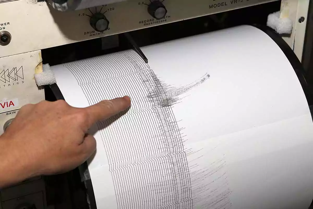 A hand points to the record of a seismograph showing a sudden increase in the lines, indicating seismic activity. A hand points to the record of a seismograph showing a sudden increase in the lines, indicating seismic activity.
