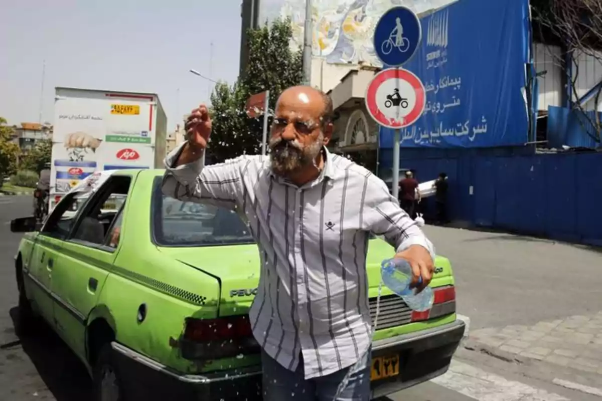 Hombre con barba y camisa de rayas se refresca con agua junto a un taxi verde en una calle urbana