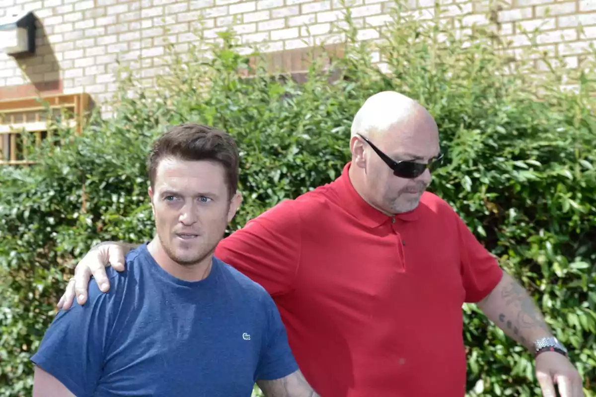 Two men walking together outdoors, one wearing a blue t-jersey and the other wearing a red polo jersey and sunglasses. Two men walking together outdoors, one wearing a blue t-jersey and the other wearing a red polo jersey and sunglasses.