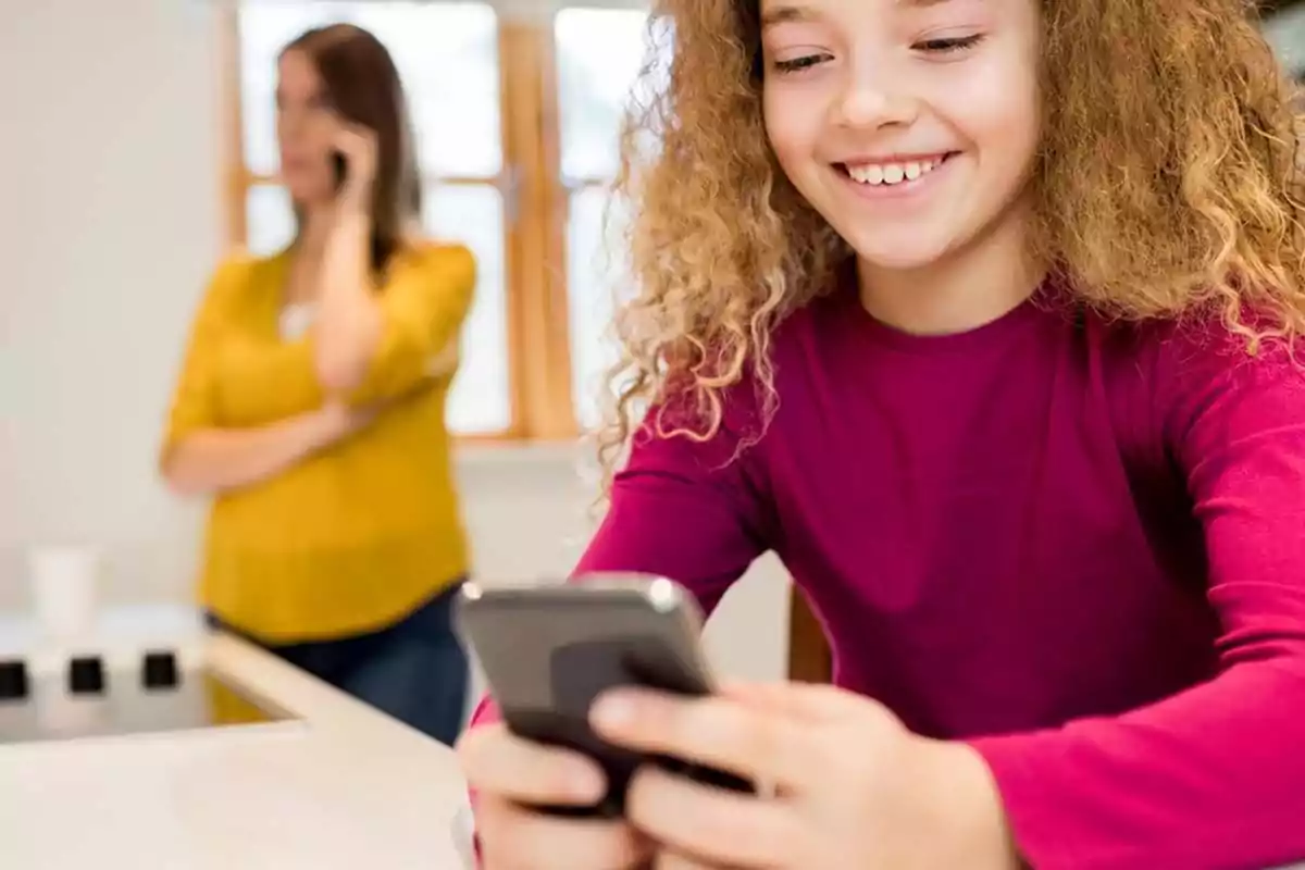 Smiling girl using a mobile phone while a woman in the background talks on the phone in a brightly lit kitchen. Smiling girl using a mobile phone while a woman in the background talks on the phone in a brightly lit kitchen.