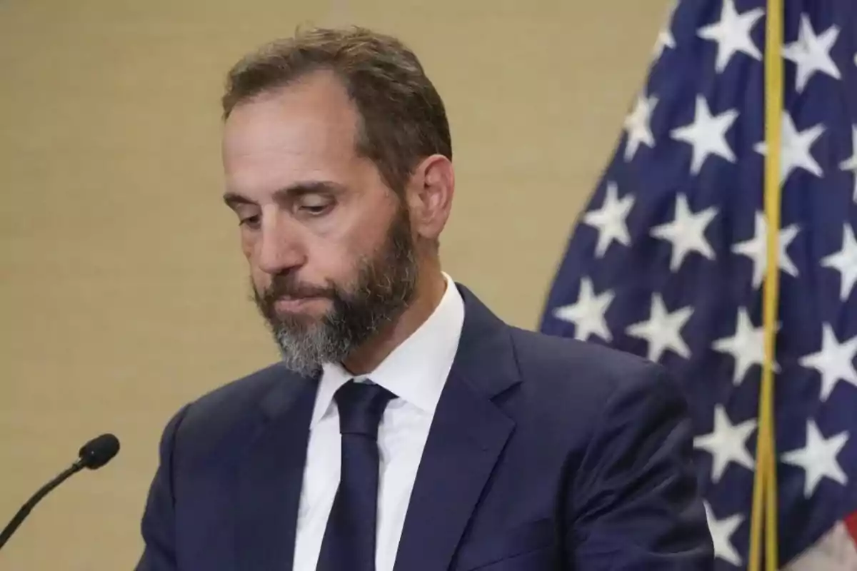 Hombre de barba y traje azul mirando hacia abajo con una bandera de Estados Unidos al fondo