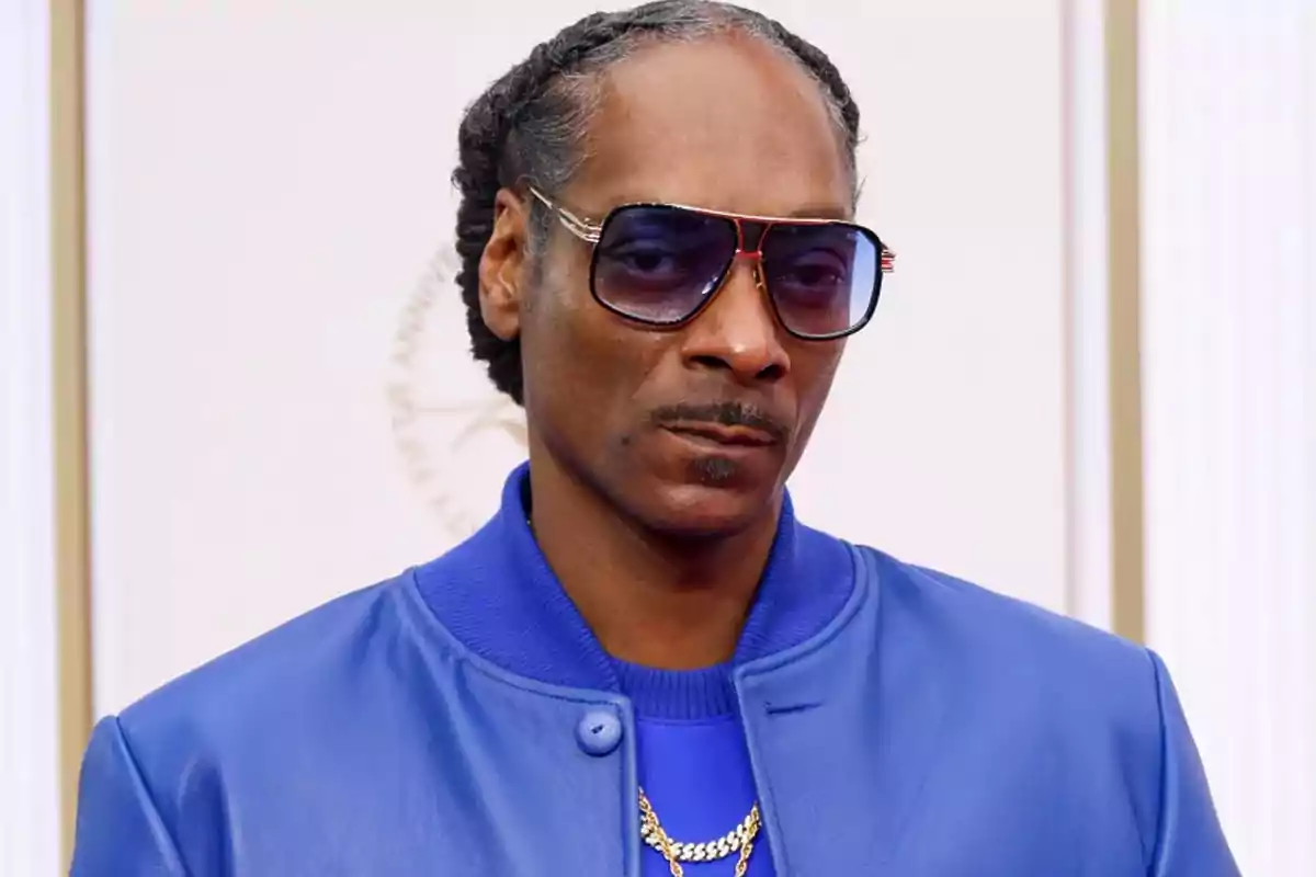 Person wearing a blue jacket, blue t-jersey, dark sunglasses, and a gold necklace, posing in front of a light background. Person wearing a blue jacket, blue t-jersey, dark sunglasses, and a gold necklace, posing in front of a light background.