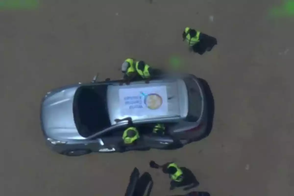Aerial view of a silver car surrounded by several people wearing reflective vests, with a sign on the roof featuring a logo and text.