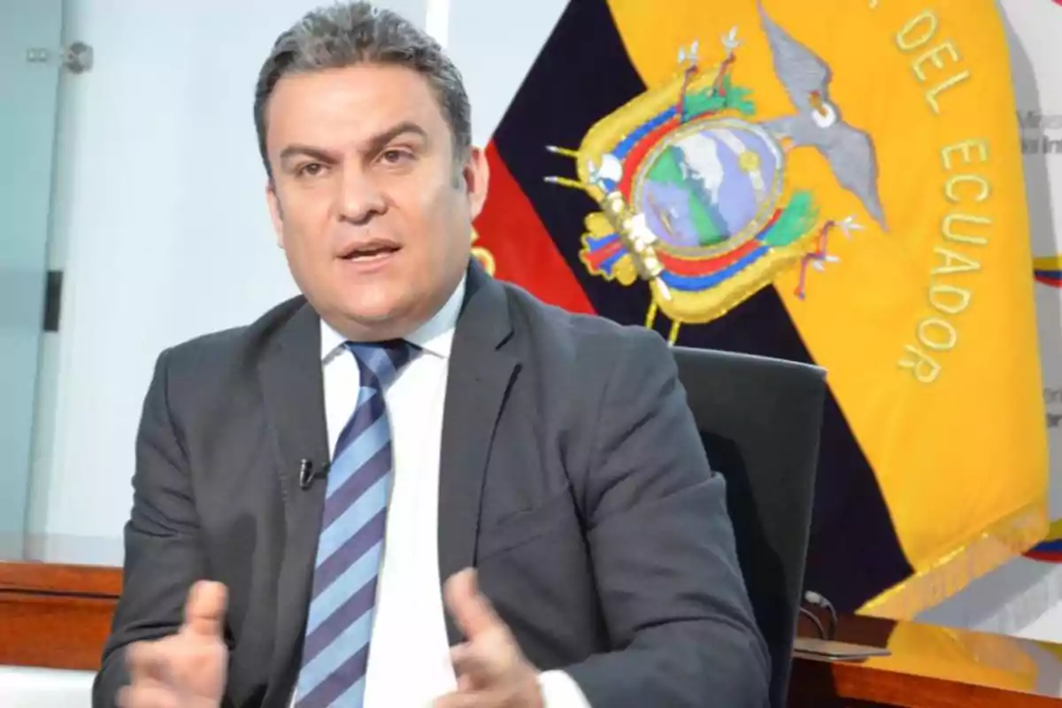 Hombre de traje hablando mientras está sentado con una bandera de Ecuador al fondo