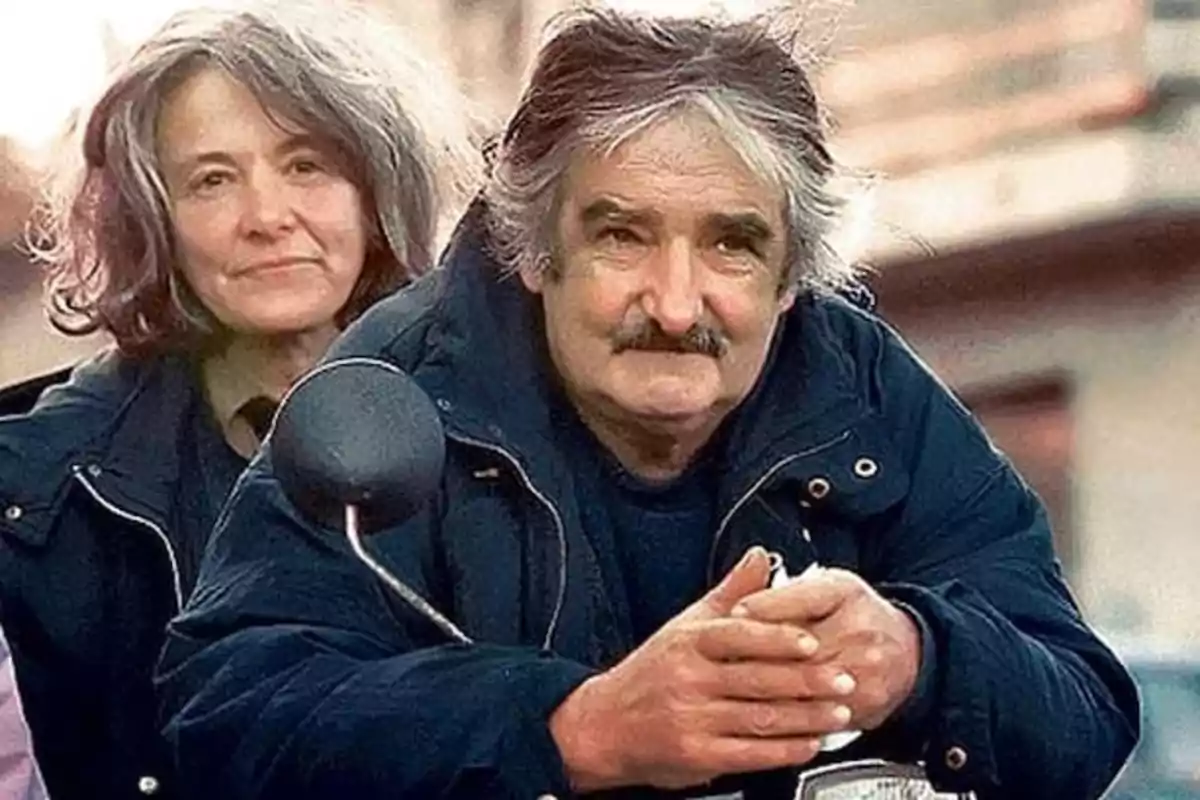 An elderly couple with gray hair and warm clothing poses on a motorcycle in a blurred urban setting. An elderly couple with gray hair and warm clothing poses on a motorcycle in a blurred urban setting.
