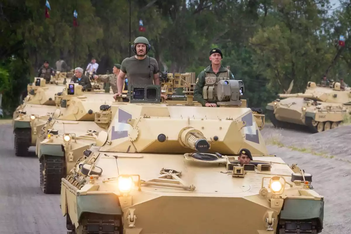 Varios tanques militares de color arena avanzan en fila por una carretera con soldados a bordo y árboles al fondo. Varios tanques militares de color arena avanzan en fila por una carretera con soldados a bordo y árboles al fondo.