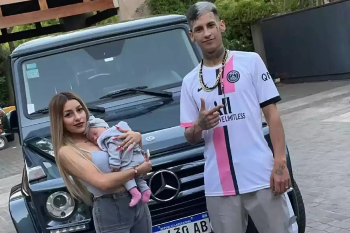 Una mujer sostiene a un bebé en brazos mientras un hombre posa a su lado frente a una camioneta Mercedes-Benz negra.