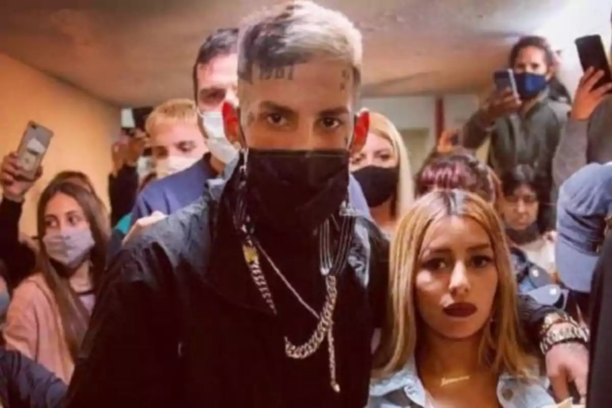 Un hombre con cabello corto y teñido de rubio, usando mascarilla negra y cadenas, posa junto a una mujer de cabello rubio y labios oscuros en un lugar cerrado rodeados de varias personas que también llevan mascarilla
