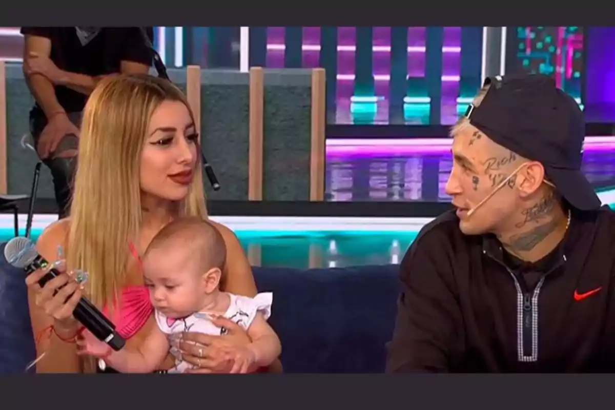 Una mujer con cabello rubio sostiene a un bebé en brazos mientras sostiene un micrófono y mira a un hombre con tatuajes en el rostro y gorra negra, ambos sentados en un set de televisión con luces de colores al fondo Una mujer con cabello rubio sostiene a un bebé en brazos mientras sostiene un micrófono y mira a un hombre con tatuajes en el rostro y gorra negra, ambos sentados en un set de televisión con luces de colores al fondo