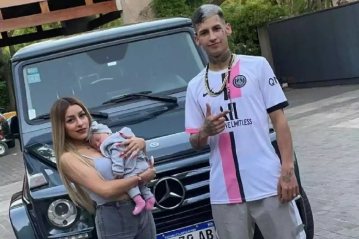 Una mujer sostiene a un bebé en brazos mientras un hombre posa a su lado frente a una camioneta Mercedes-Benz negra. Una mujer sostiene a un bebé en brazos mientras un hombre posa a su lado frente a una camioneta Mercedes-Benz negra.
