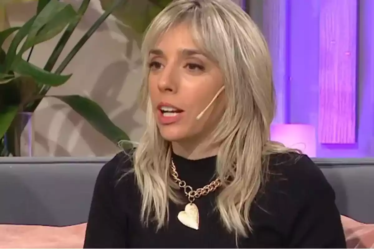 Blonde woman with a headset microphone sitting on a sofa during an interview, wearing a gold necklace with a heart pendant and a black sweater, background with plants and purple lights.