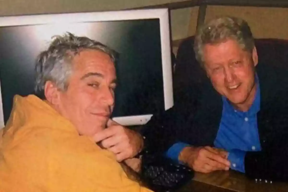 Two men sitting together in front of a computer monitor, both looking at the camera and smiling.
