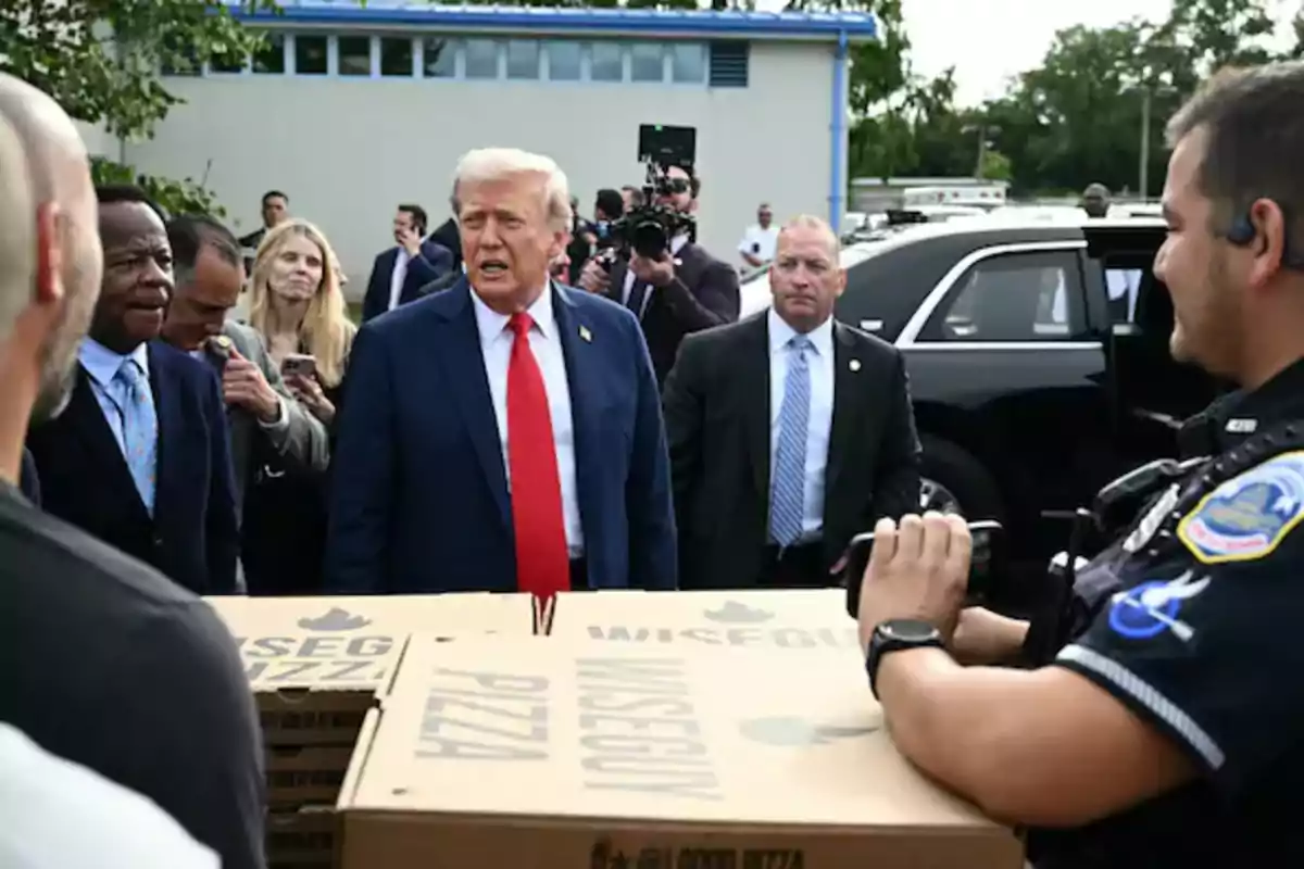 A man in a blue suit and red tie is surrounded by several people, including a police officer holding pizza boxes, while a camera records the scene outdoors next to a black car.