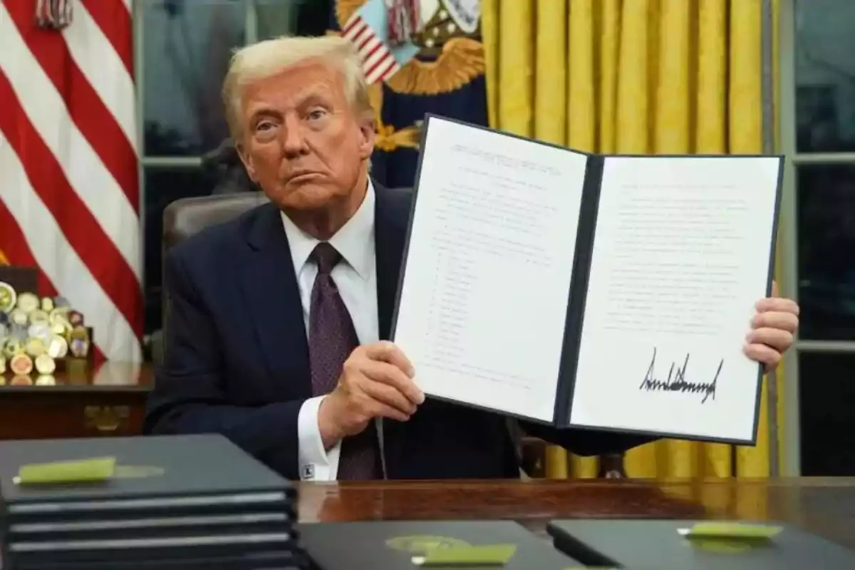 Man in a suit sitting at a desk holding an open document with a visible signature, with yellow curtains and a United States flag in the background. Man in a suit sitting at a desk holding an open document with a visible signature, with yellow curtains and a United States flag in the background.