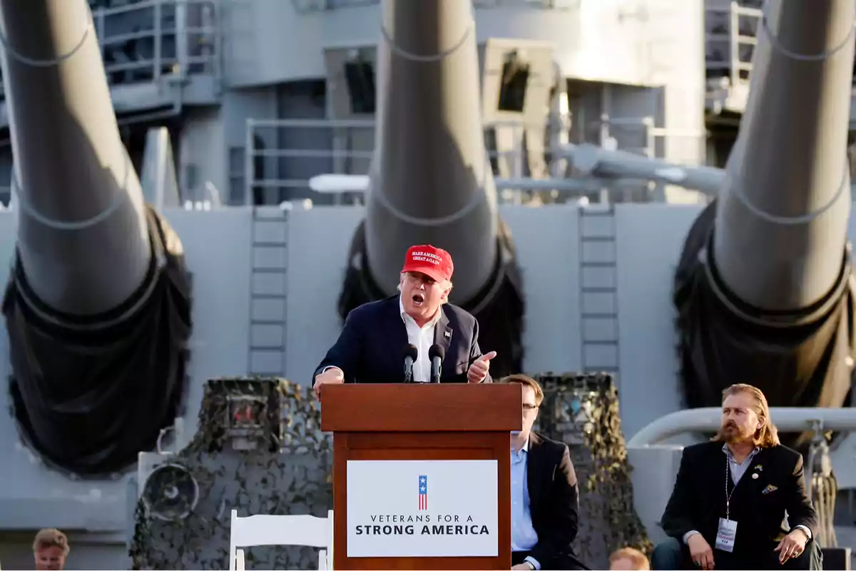 Man with a red cap speaking at a podium in front of a military ship accompanied by other people Man with a red cap speaking at a podium in front of a military ship accompanied by other people